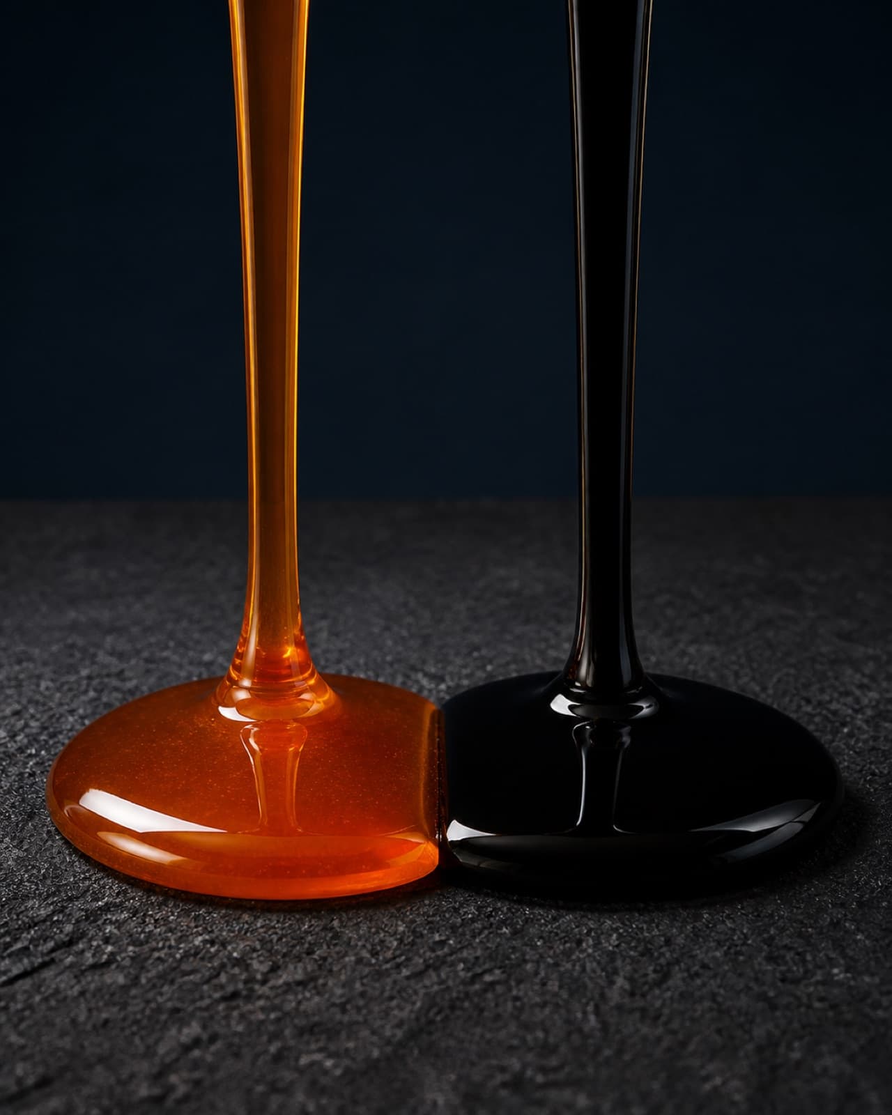 Two liquids poured side by side onto a dark textured surface, one amber and one black, meeting at the edge