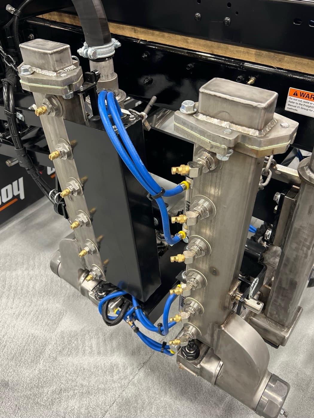 Two folded stainless spray bar sections with nozzles and blue supply lines on the side of a LeeBoy distributor