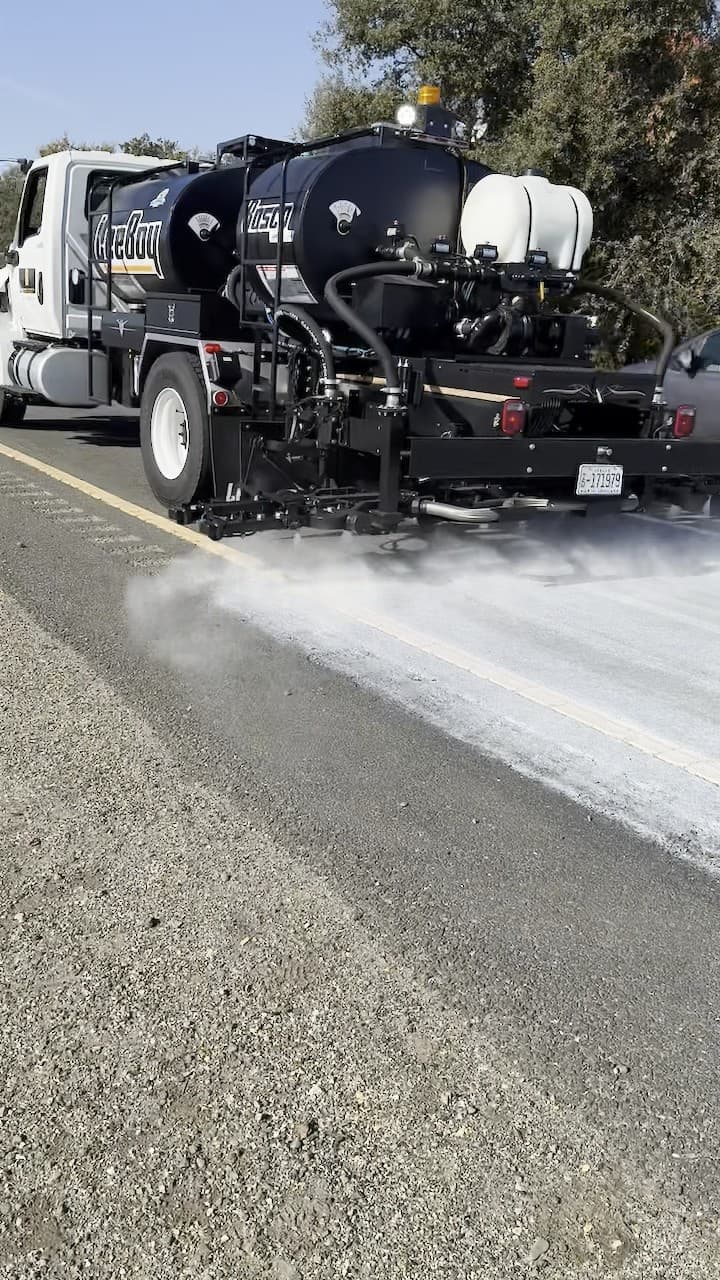Distributor truck with two tanks spraying a highway lane from its rear spray bar, freshly treated surface behind it