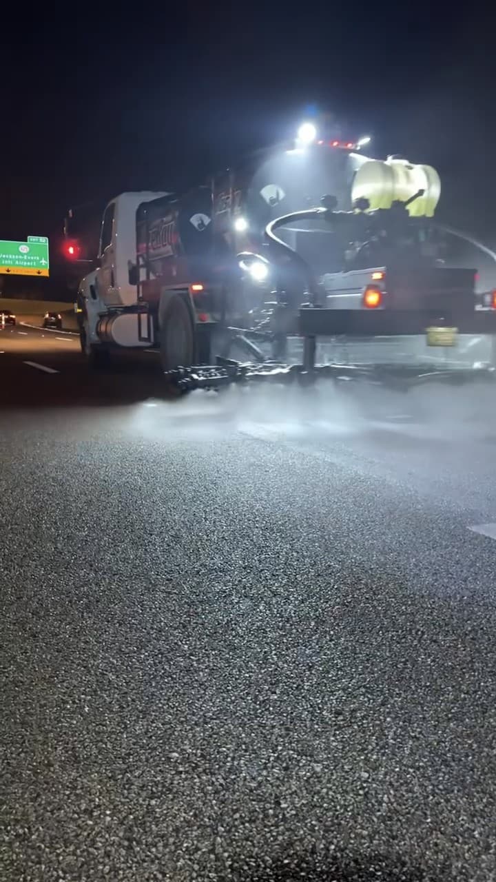Distributor truck spraying a highway lane at night under its work lights, with traffic headlights beyond