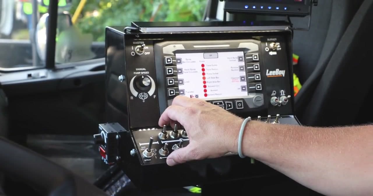 Operator's hand at the in-cab control console of a LeeBoy PR900 distributor, a color display above a row of toggle switches