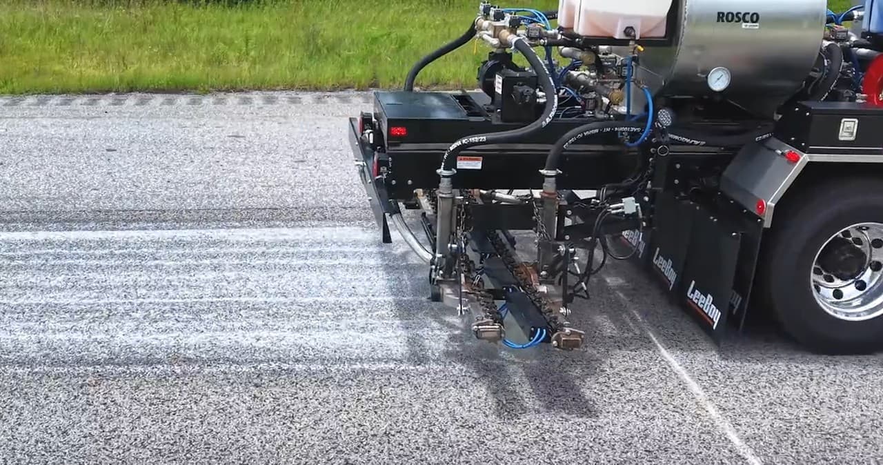 Rear spray bar of a LeeBoy PR900 distributor seen from the side, nozzles spraying onto a lined test lane