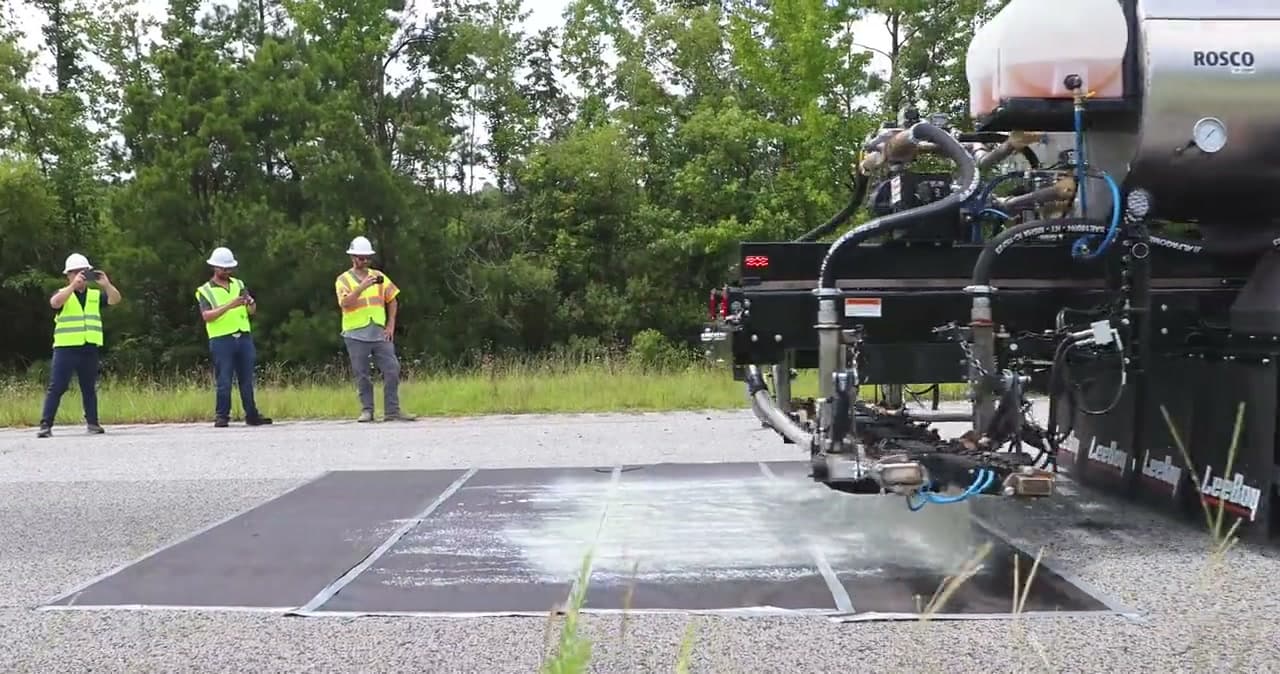 LeeBoy PR900 distributor spraying a marked test section on a paved lot, crew standing clear in the background